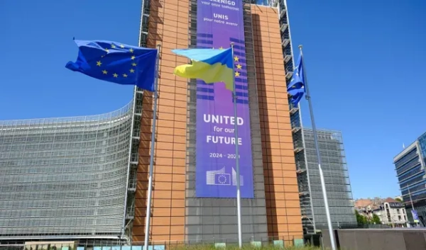 Прапори ЄС та України майорять на вітрі перед будівлею Берлемон, штаб-квартирою Європейської Комісії, з нагоди Дня Незалежності України, 24 серпня 2025 року в Брюсселі, Бельгія. На фасаді висить великий банер з написом англійською та французькою мовами: