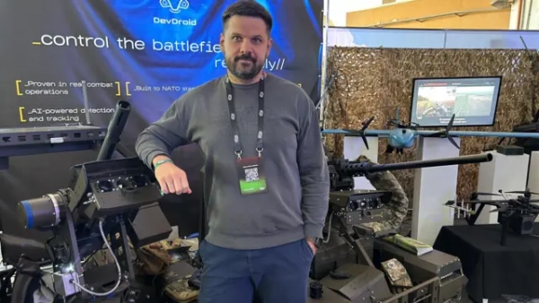 Автор фото, Serhiy Beskrestnov Вадим, чоловік, якому на вигляд близько сорока років, стоїть за двома кулеметами. Кулемети розміщені на платформах і мають чорний колір. Позаду нього - стенд, що пояснює їхні властивості, і модель літака.