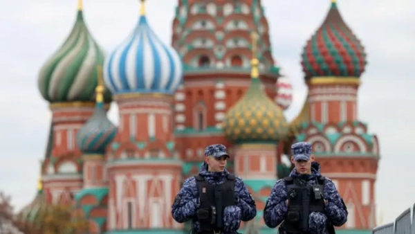Двоє чоловіків у синьо-чорній камуфляжній уніформі, кепках і чорних жилетах розмовляють, прямуючи повз червоний кам’яний собор із блакитними, зеленими, червоними та золотими оздобами на куполах.
