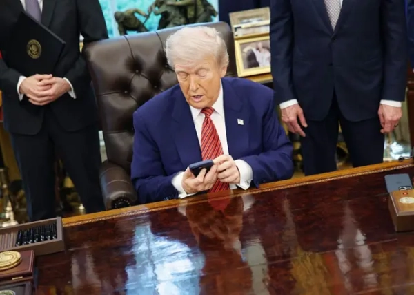 Трамп за столом в Овальному кабінеті тримає обома руками смартфон, сидячи в шкіряному кріслі. По обидва боки від нього – чоловіки в костюмах, їхні обличчя поза кадром