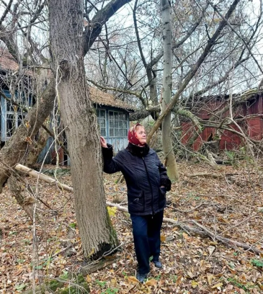 Автор фото, Державне агентство з управління зоною відчуження Жінка у чорній куртці, широких джинсах і червоній хустці стоїть серед занедбаного подвір’я, опершись на стовбур дерева. Навколо багато сухого пожовклого листя та гілок, а в тлі видно покинутий дерев’яний будинок.