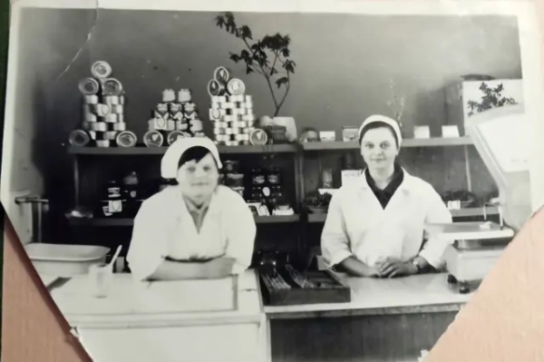 Автор фото, Державне агентство з управління зоною відчуження Чорно-біле фото, зроблене всередині магазину або кулінарного відділу. На передньому плані – прилавок з механічними вагами. За прилавком стоять двоє жінок у білих халатах і головних уборах. На задньому фоні – дерев’яні полиці, заповнені товарами: піраміди консервних банок, скляні банки з продуктами, кілька коробок