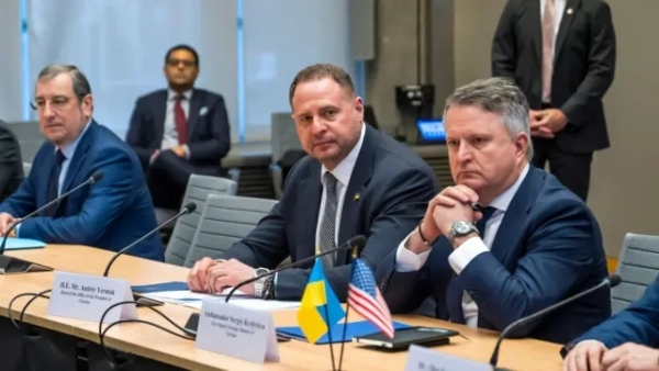 На світлині кілька чоловіків у діловому вбранні сидять за великим столом для переговорів. На передньому плані Єрмак, котрий дивиться майже в об'єктив, і Кислиця, що сидить зі складеними перед обличчям руками, сконцентрований. На столі перед ними знаходяться невеличкі прапори України та Сполучених Штатів. На задньому плані видно інших учасників зустрічі й охоронця, котрий стоїть біля стіни.