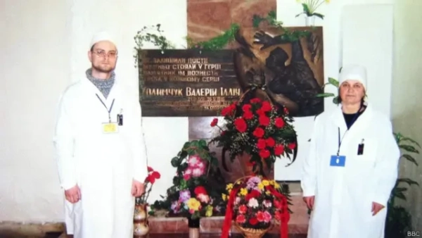 Автор фото, Державне агентство з управління зоною відчуження Чоловік і жінка у білих халатах та шапочках стоять по обидва боки меморіальної плити, уквітчаної великими букетами червоних, рожевих і жовтих квітів. На плиті – рельєфна скульптурна композиція та напис українською мовою: "Не залишили пости, мужньо стояли у герці, памʼятник їм вознести треба у кожному серці. Ходимчук Валерій Ілліч"