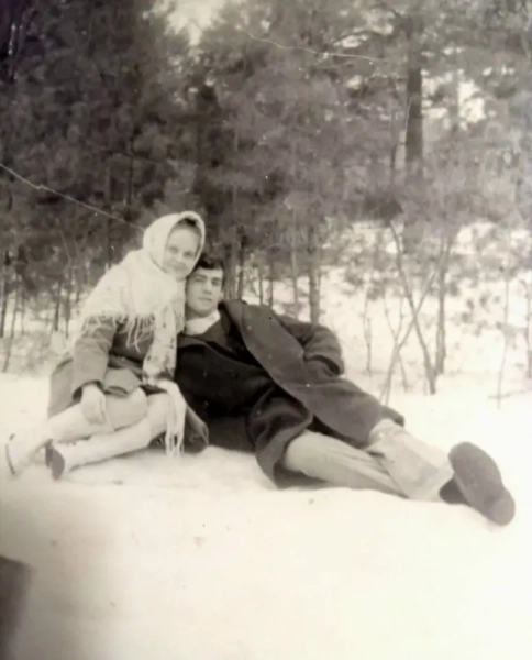 Автор фото, Державне агентство з управління зоною відчуження На чорно-білому знімку молоде подружжя сидить разом на снігу серед зимового лісу. Жінка в хустці та теплому пальті сидить збоку й ніжно схиляється до чоловіка. Чоловік у темному пальті напівлежить поруч, водночас трохи обіймаючи її. Обоє виглядають спокійними та близькими, позують на фоні високих засніжених дерев