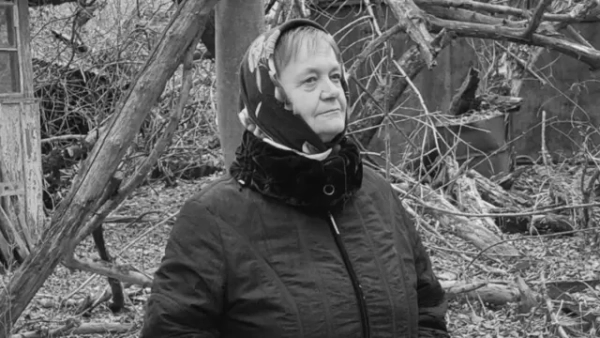 Автор фото, Державне агентство з управління зоною відчуження Чорно-білий портрет літньої жінки в куртці та хустці з квітами на тлі повалених дерев покинутого двору