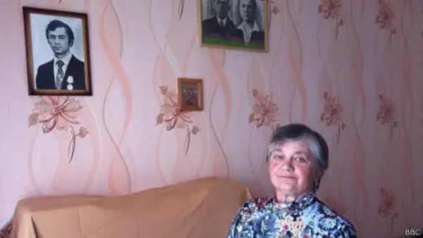 Автор фото, Державне агентство з управління зоною відчуження На світлині літня жінка сидить у кімнаті з рожевими шпалерами, прикрашеними квітковим візерунком. Вона усміхається м’якою, спокійною усмішкою. Позаду неї на стіні висять три портрети: великий чорно-білий портрет молодого чоловіка в костюмі з медалями, поруч – менший кольоровий портрет подружжя та маленька іконка в рамці/